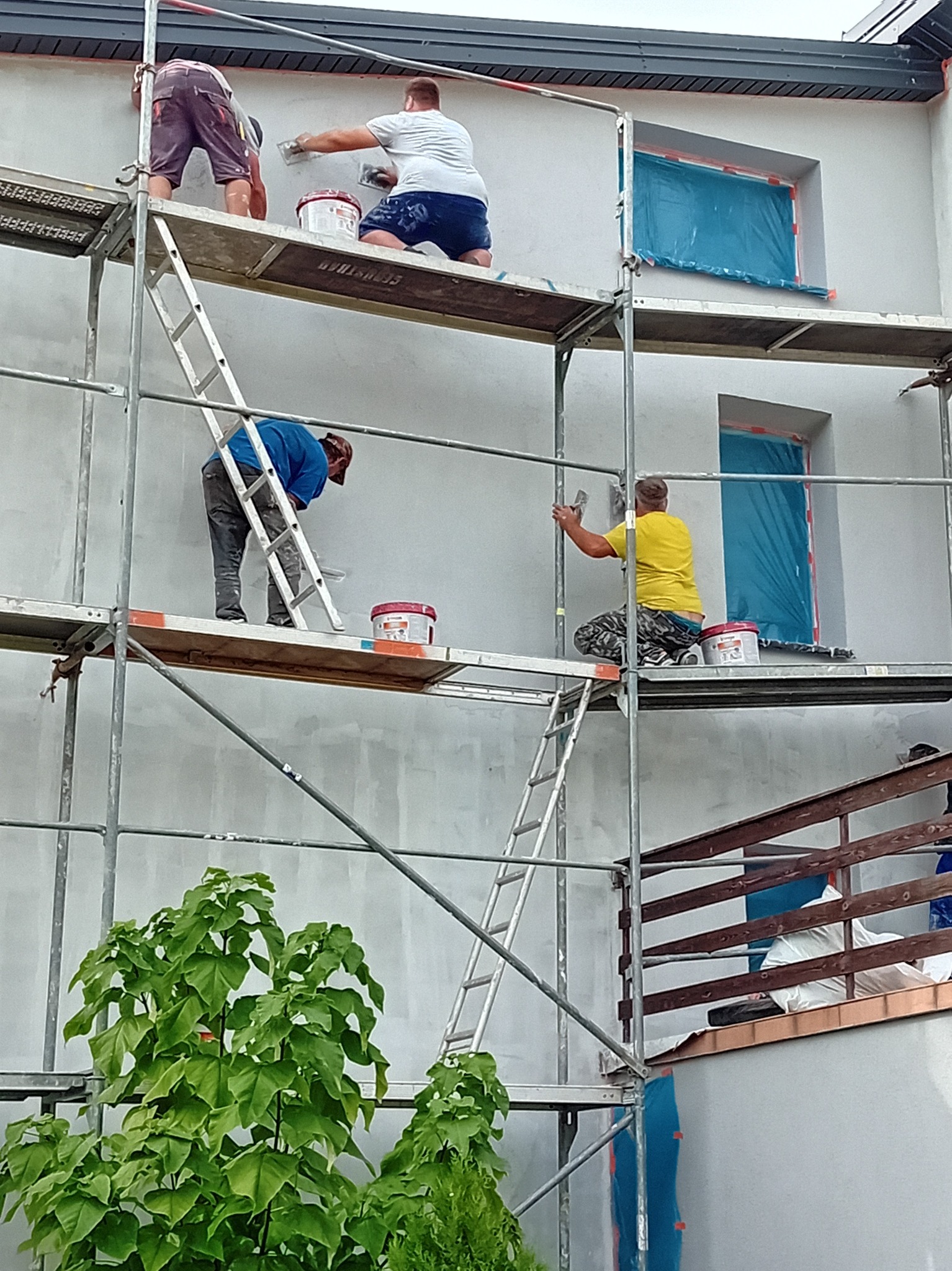 Czterech mężczyzn na rusztowaniu nakłada zaprawę na elewację budynku, okna zabezpieczone folią, widoczne wiadra z zaprawą.
