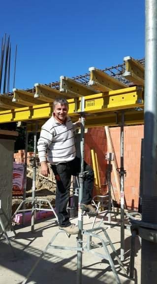 Uśmiechnięty mężczyzna stoi na rusztowaniu pod żółtymi belkami szalunkowymi, przygotowanymi do zalania betonem, widoczne zbrojenia i ceglany mur w tle.