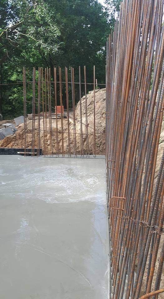 Świeżo wylany beton z widocznym zbrojeniem pionowym i poziomym, przygotowany pod fundament, w tle ziemia i drzewa.