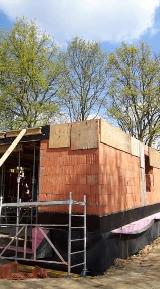 Budowa domu jednorodzinnego: ściany z czerwonej cegły, widoczne deskowanie na górze, izolacja fundamentów z czarnej folii i różowego styropianu, rusztowanie przy ścianie, drzewa w tle.