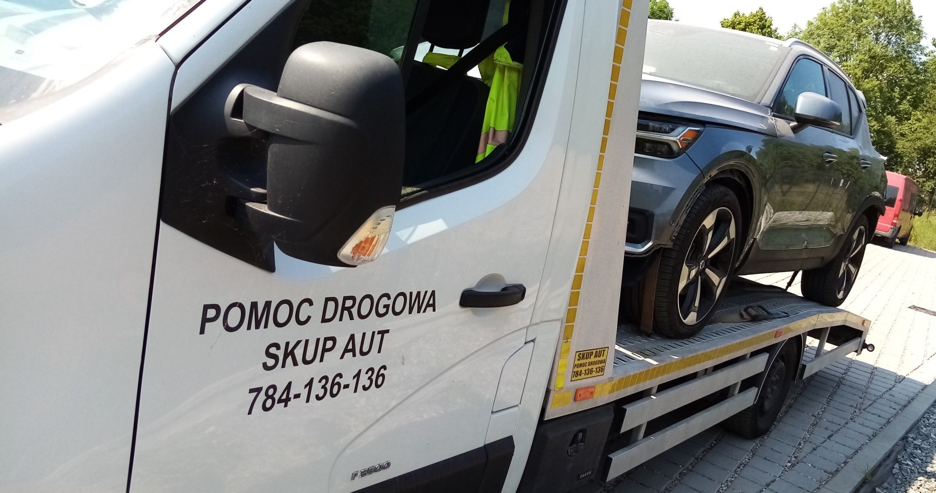 Szary samochód osobowy przewożony na lawecie z napisem 'Pomoc Drogowa Skup Aut 784-136-136' na białej kabinie.