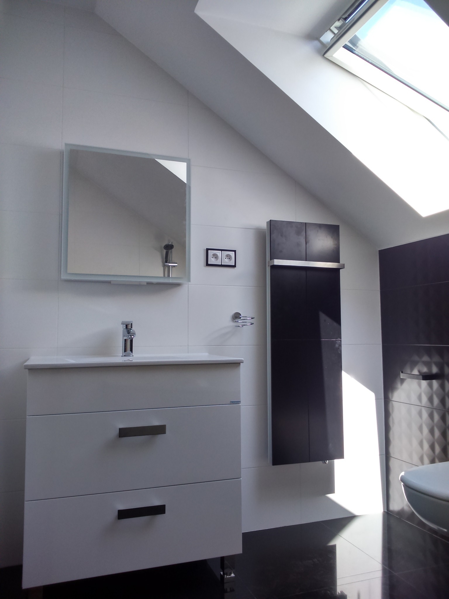 Nowoczesna łazienka na poddaszu z białymi płytkami, szafką z umywalką i lustrem, czarnym grzejnikiem drabinkowym oraz oknem dachowym wpuszczającym jasne światło.