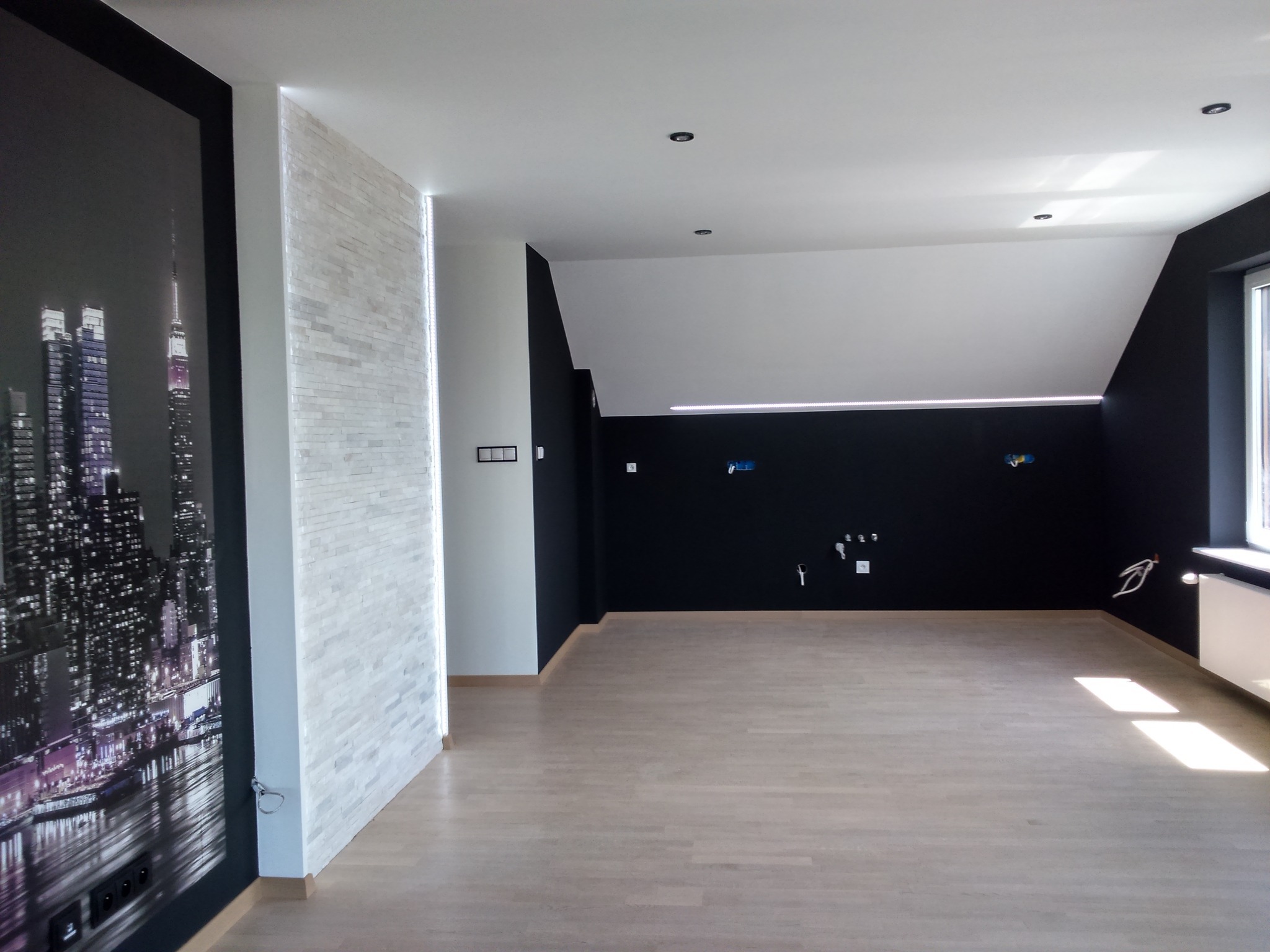 Nowoczesne wnętrze poddasza po remoncie: czarna ściana, jasne panele podłogowe, fototapeta z motywem miasta i dekoracyjna ściana z podświetleniem LED.