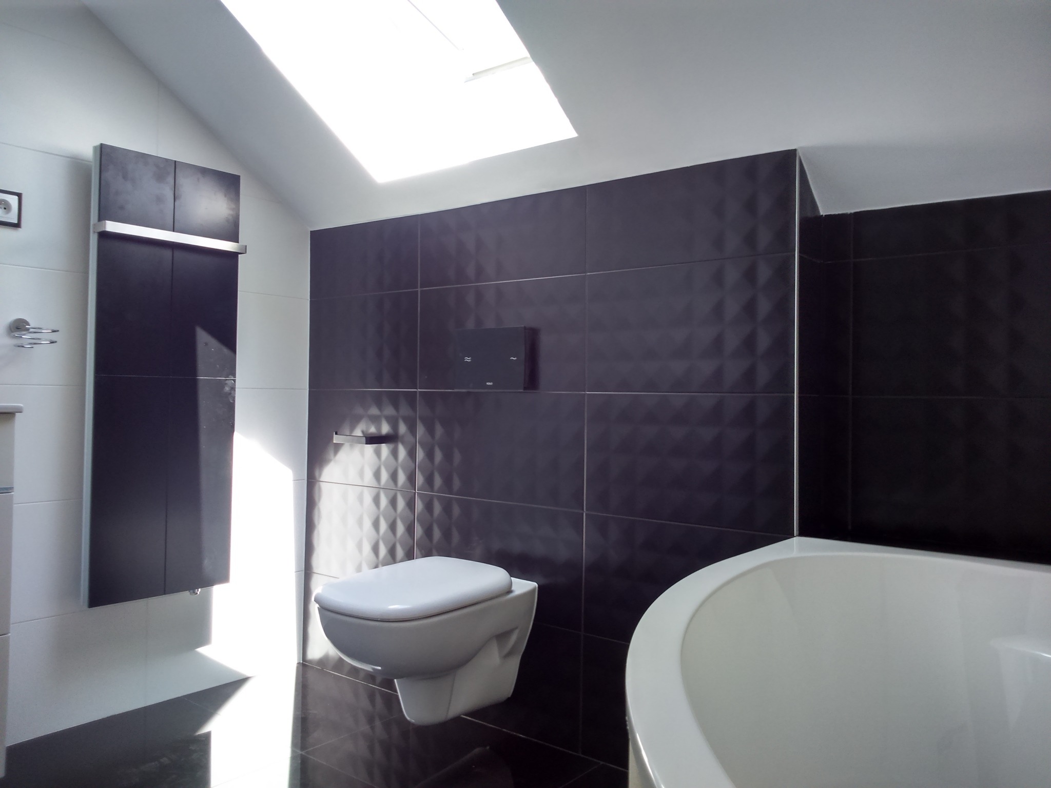 Nowoczesna łazienka na poddaszu z czarnymi, trójwymiarowymi płytkami na ścianie, białą armaturą i grzejnikiem panelowym, oświetlona światłem z okna dachowego.