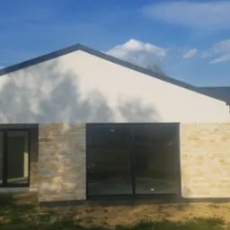 Nowoczesny dom parterowy z białymi ścianami, fragmentami elewacji z kamienia naturalnego oraz dużymi przesuwnymi oknami z czarnymi ramami, drewniane panele dekoracyjne, dach w kolorze antracytowym.