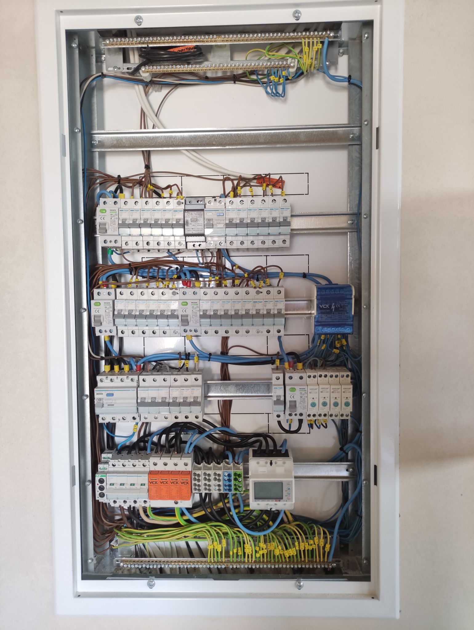 Rozdzielnica elektryczna z zabezpieczeniami, przewodami w kolorach: niebieskim, brązowym, żółto-zielonym. Zamontowana w białej obudowie.