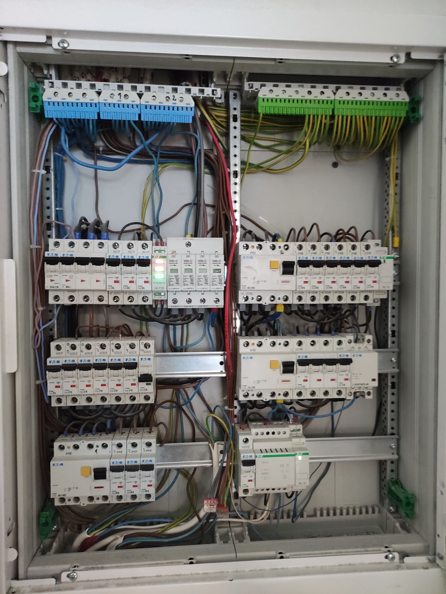 Rozdzielnica elektryczna w obudowie, widoczne liczne przewody, bezpieczniki i listwy zaciskowe. Elementy instalacji w kolorach: niebieskim, zielonym, żółtym i czerwonym.