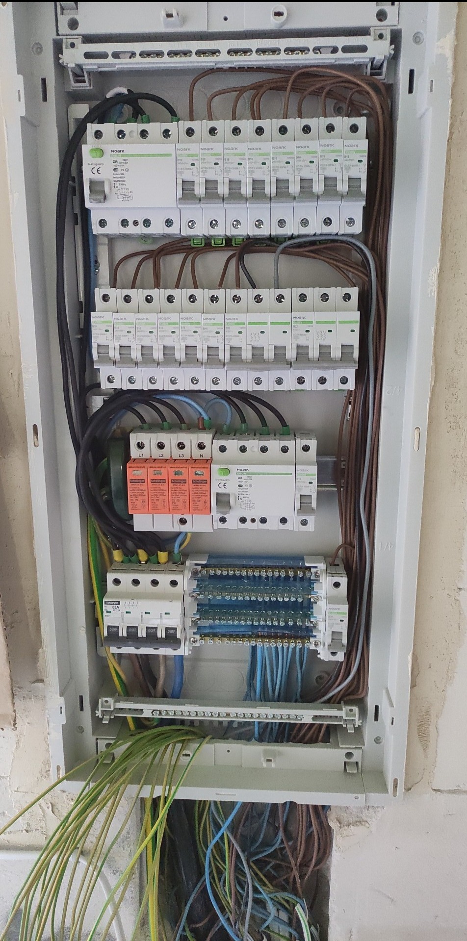 Rozdzielnica elektryczna w trakcie montażu, widoczne zabezpieczenia, przewody i listwy zaciskowe. Kable w różnych kolorach. Biała obudowa rozdzielnicy.