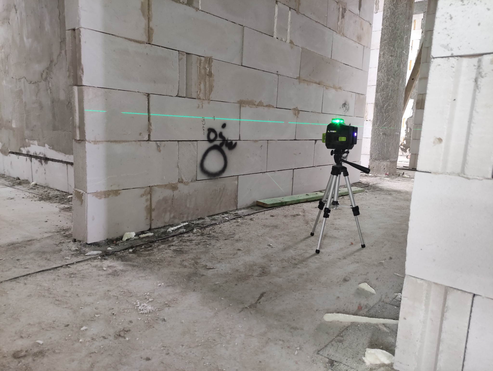 Laser krzyżowy na statywie wyznacza poziom na ścianie z betonu komórkowego z graffitti. Widoczny fragment instalacji elektrycznej i surowy stan wykończenia.