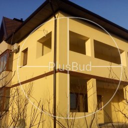 PLUSBUD - Żółta elewacja dwupiętrowego domu z brązowymi oknami i dachem, widoczny klimatyzator na ścianie, nałożone logo firmy PlusBud.