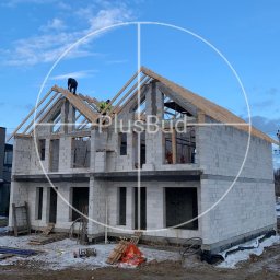 PLUSBUD - Wznoszenie dwukondygnacyjnego domu z bloczków betonowych, widoczne drewniane elementy konstrukcji dachu, dwóch robotników na dachu, plac budowy z narzędziami i materiałami.