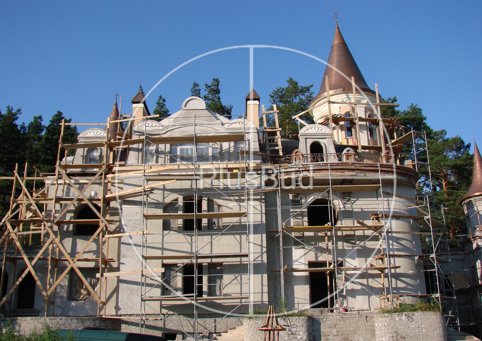 Budowa zamku w stylu baśniowym, widoczne rusztowania i surowe ściany z elementami dekoracyjnymi, wieże z miedzianymi dachami w otoczeniu drzew.