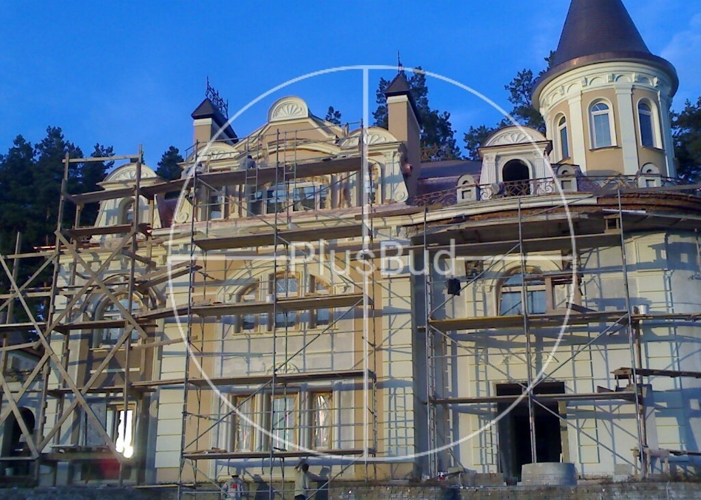 Elewacja budynku w stylu pałacowym w trakcie prac wykończeniowych, widoczne rusztowania i elementy dekoracyjne, tło z drzew iglastych i błękitne niebo.