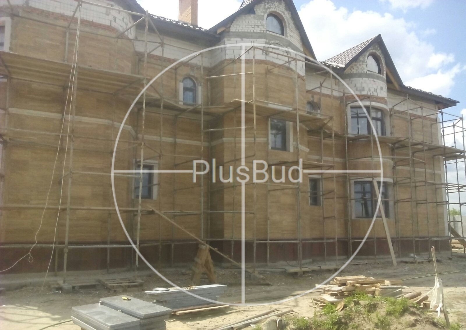 Budynek dwupiętrowy w trakcie prac elewacyjnych, widoczne rusztowania i przygotowane płyty na ziemi.