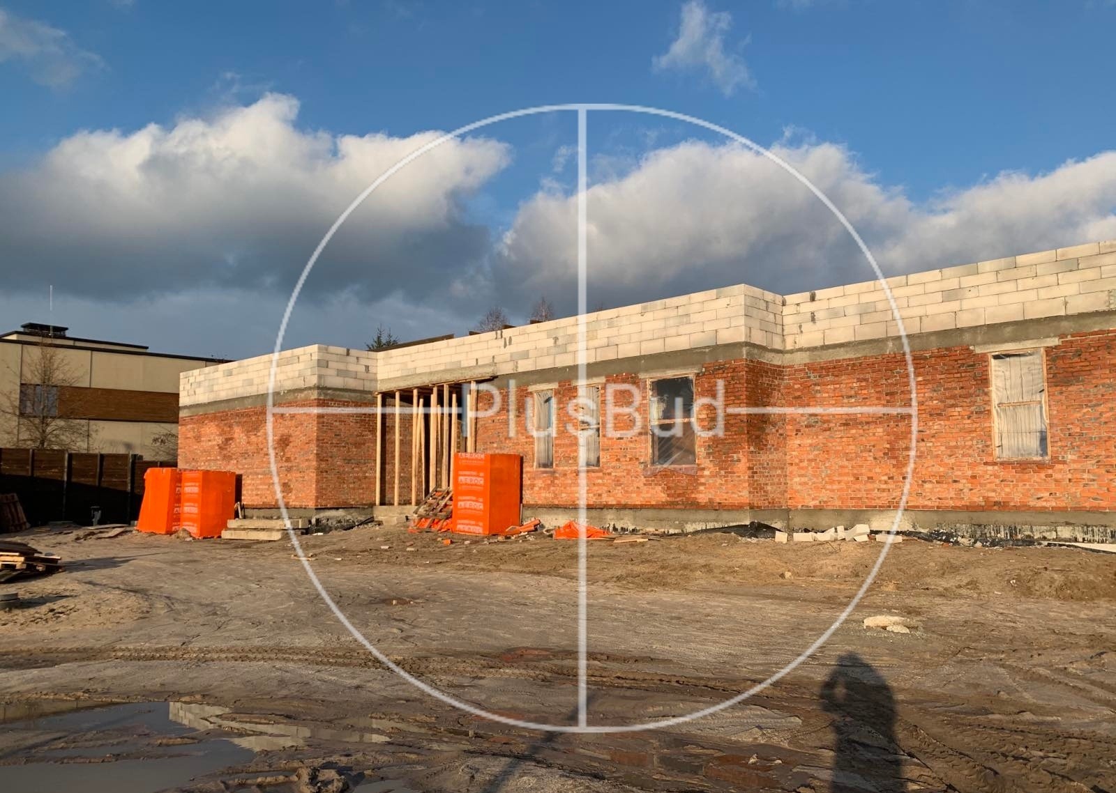 Wznoszenie ścian z czerwonej cegły i bloczków betonowych na budowie domu jednorodzinnego, widoczne pakiety materiałów budowlanych, na pierwszym planie błoto po deszczu.