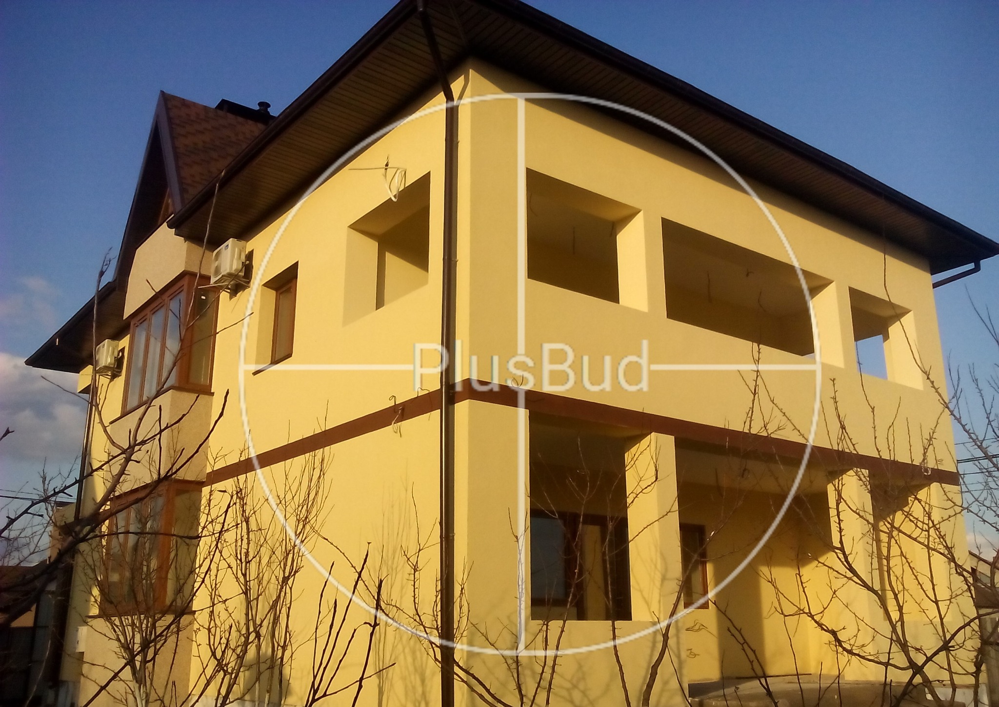 Żółta elewacja dwupiętrowego domu z brązowymi oknami i dachem, widoczny klimatyzator na ścianie, nałożone logo firmy PlusBud.