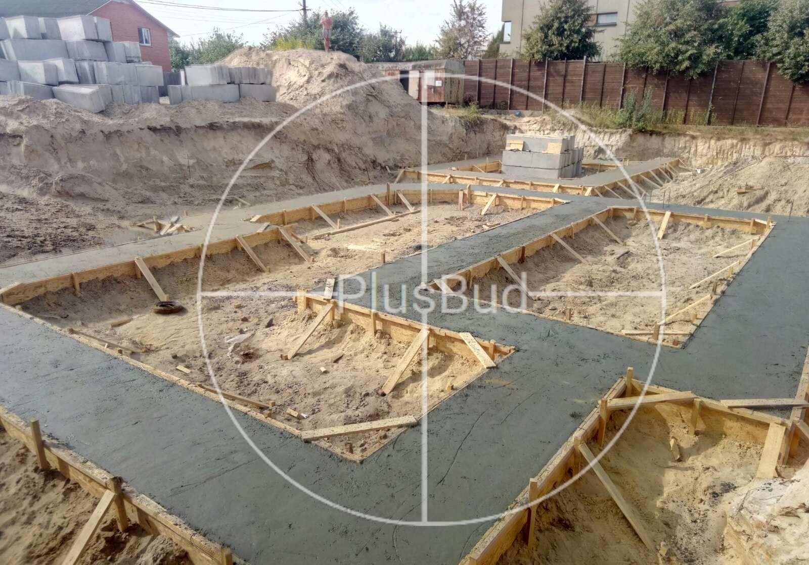 Wylane fundamenty domu w szalunkach drewnianych, widok z góry, na dalszym planie sterta piasku i bloczki betonowe.