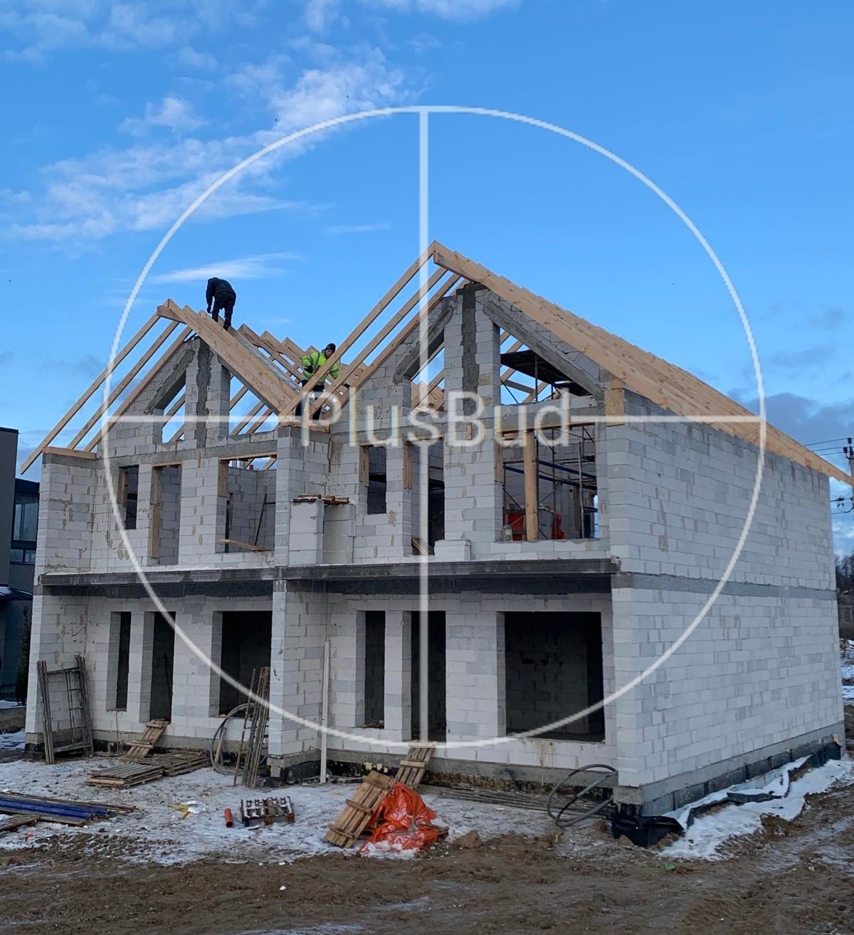 Wznoszenie dwukondygnacyjnego domu z bloczków betonowych, widoczne drewniane elementy konstrukcji dachu, dwóch robotników na dachu, plac budowy z narzędziami i materiałami.