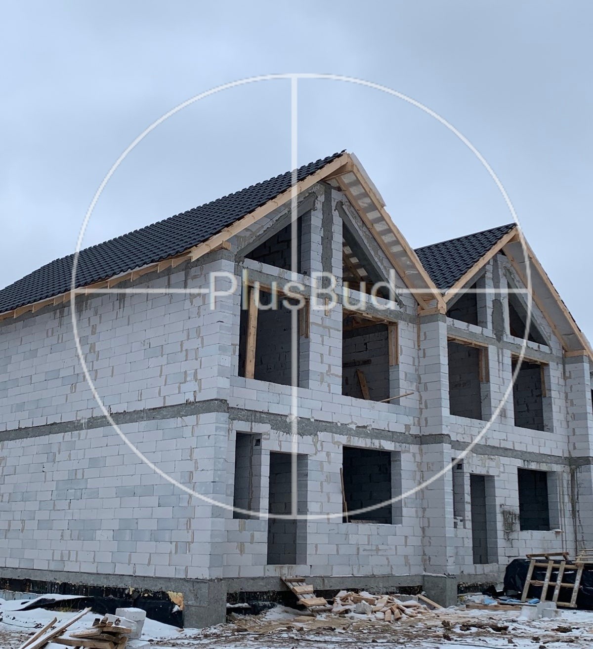 Dom w stanie surowym otwartym z białego bloczka, widoczne drewniane elementy konstrukcji dachu i czarna dachówka, na ziemi resztki śniegu i materiały budowlane.