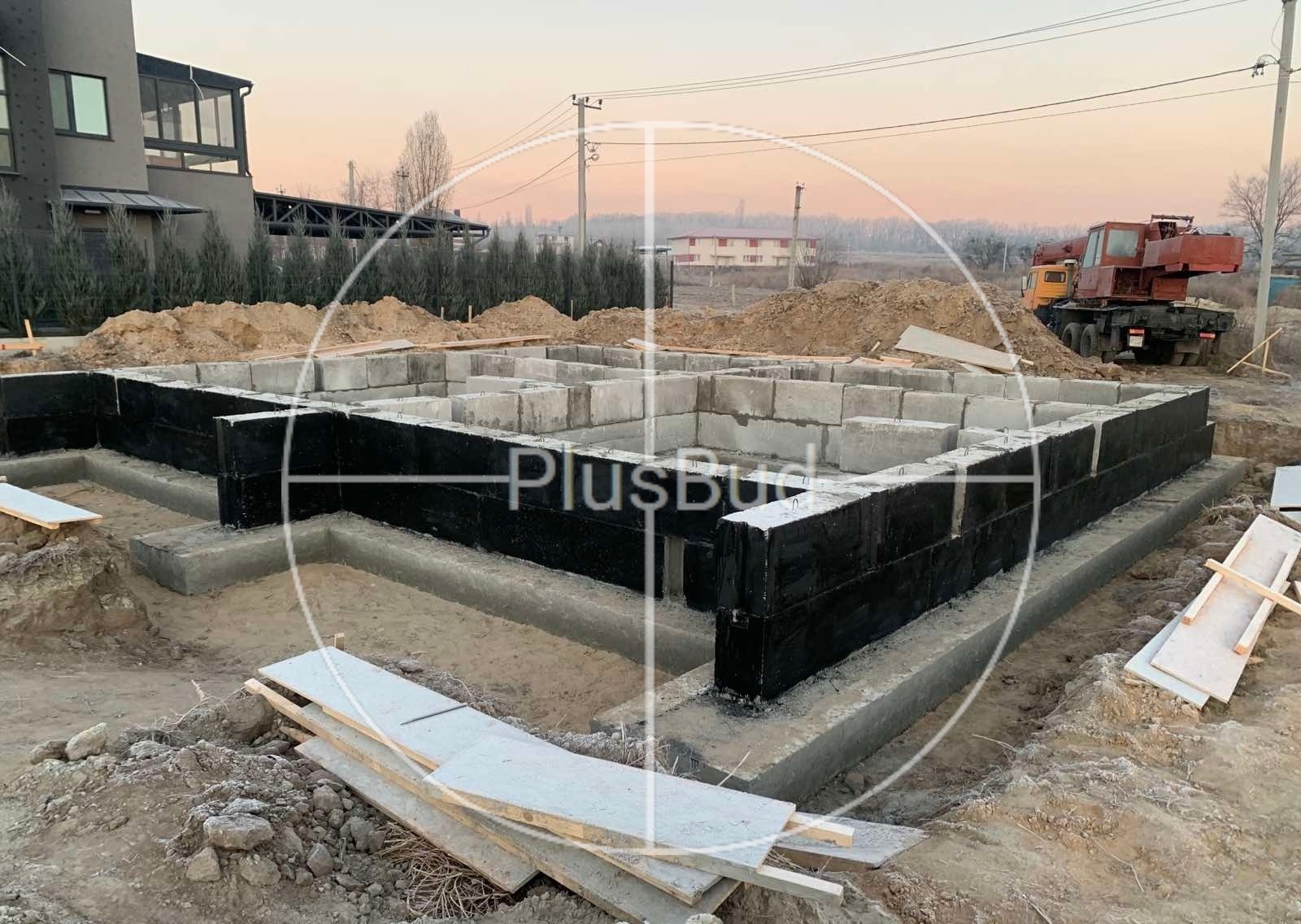 Wykopane fundamenty domu z izolacją pionową, widoczny dźwig w tle, teren budowy.