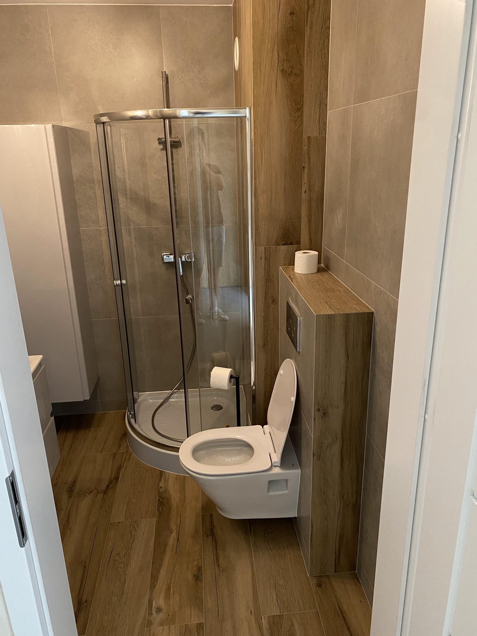 Nowoczesna łazienka z kabiną prysznicową z zaokrąglonym szkłem, podwieszaną toaletą w zabudowie z drewnopodobną okleiną i płytkami w odcieniach beżu oraz podłogą imitującą drewno.
