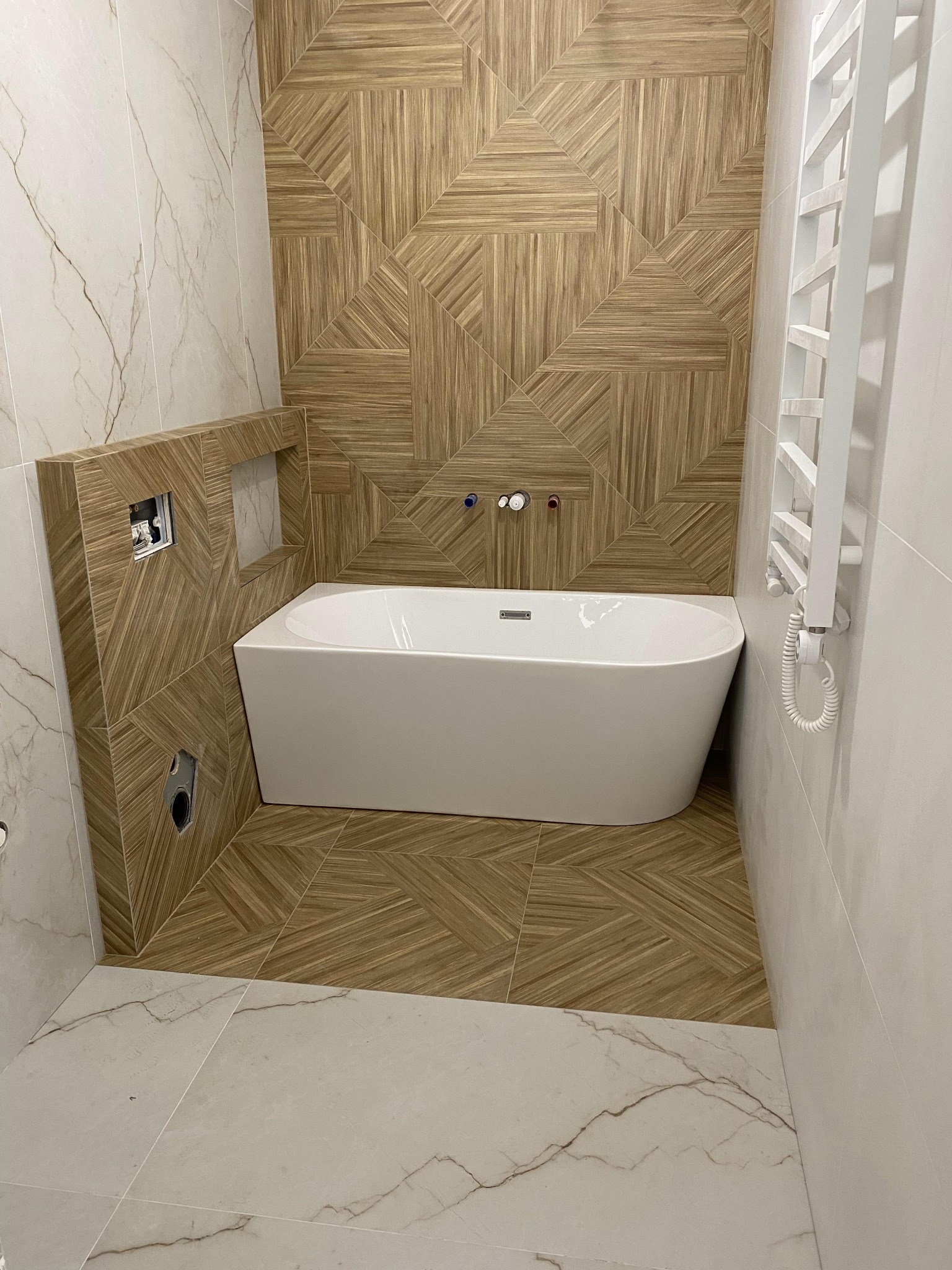 Nowoczesna łazienka z wolnostojącą wanną, ściany wyłożone płytkami imitującymi drewno ułożonymi w geometryczny wzór, białe płytki marmurowe na części ścian i podłodze, grzejnik drabinkowy.