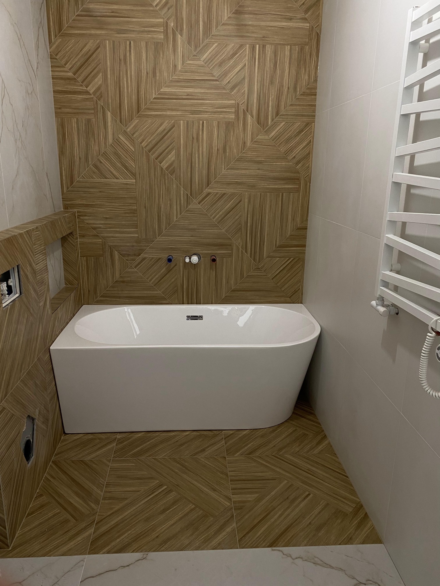 Nowoczesna łazienka z białą wanną w zabudowie z drewnopodobnych płytek ułożonych w geometryczny wzór, grzejnik drabinkowy na ścianie z jasnych płytek.