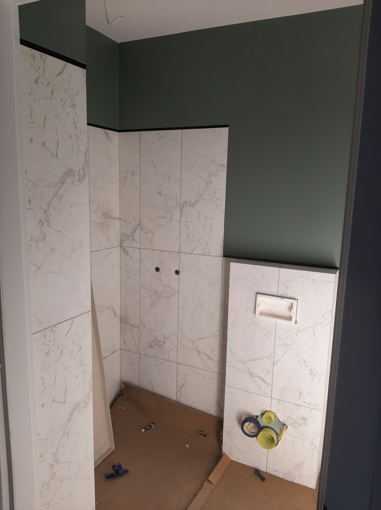 Wnętrze łazienki w trakcie wykańczania z marmurowymi płytkami na ścianach, widoczna instalacja podtynkowego systemu WC, zielona ściana nad płytkami oraz elementy instalacyjne na podłodze...