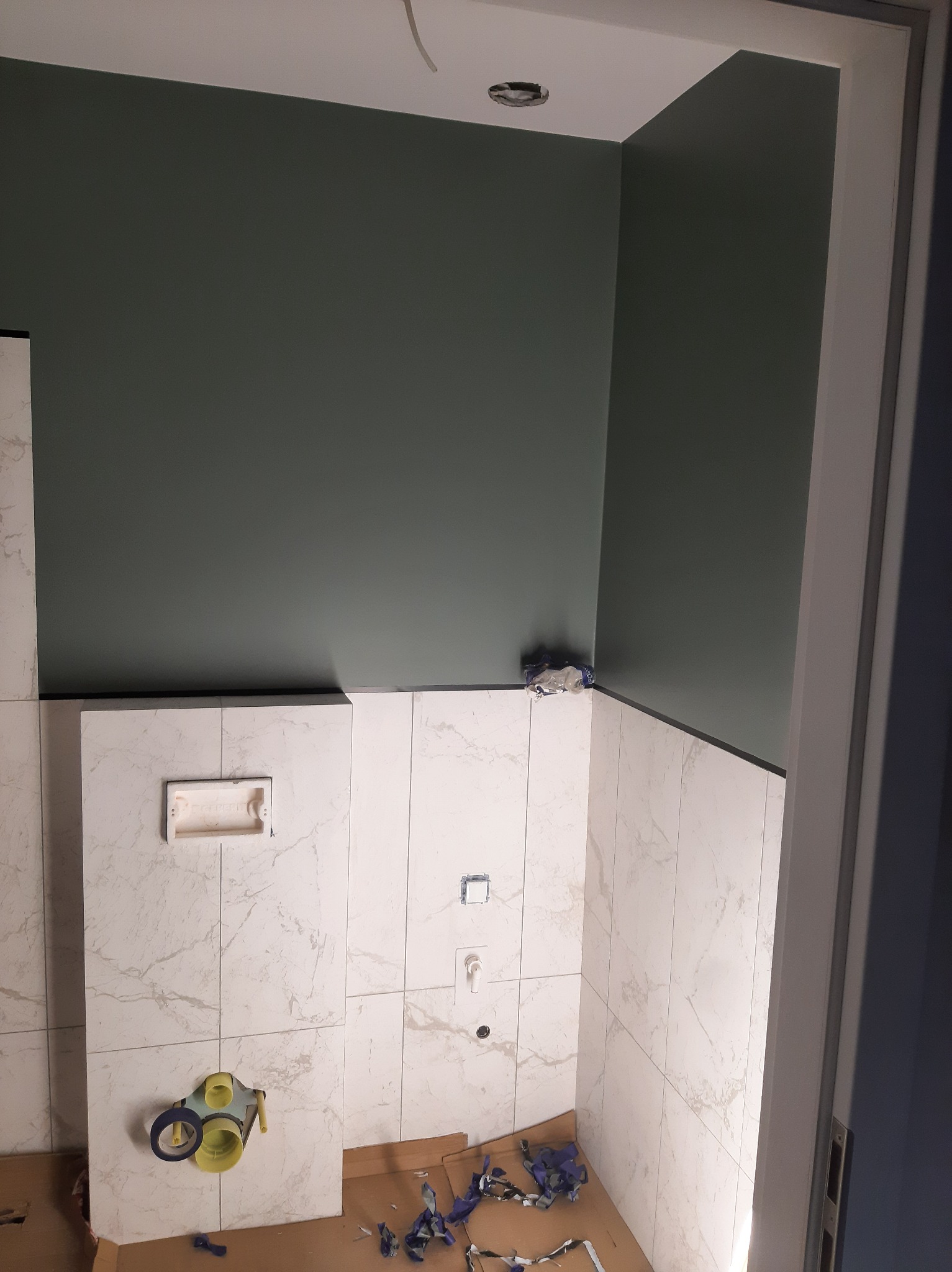 Łazienka w trakcie prac wykończeniowych: oliwkowa ściana, marmurowe płytki, widoczne elementy instalacji wodno-kanalizacyjnej i kartony na podłodze.
