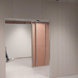 Hubert Mitręga - Wnętrze w trakcie prac wykończeniowych, widoczne ściany z płyt, częściowo zabudowane profile, instalacje pod sufitem, betonowa podłoga.