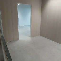 Hubert Mitręga - Surowe wnętrze: ściany wyłożone płytami gipsowo-kartonowymi, widoczna instalacja przeciwpożarowa pod sufitem, surowa podłoga i otwór drzwiowy do kolejnego pomieszczenia.