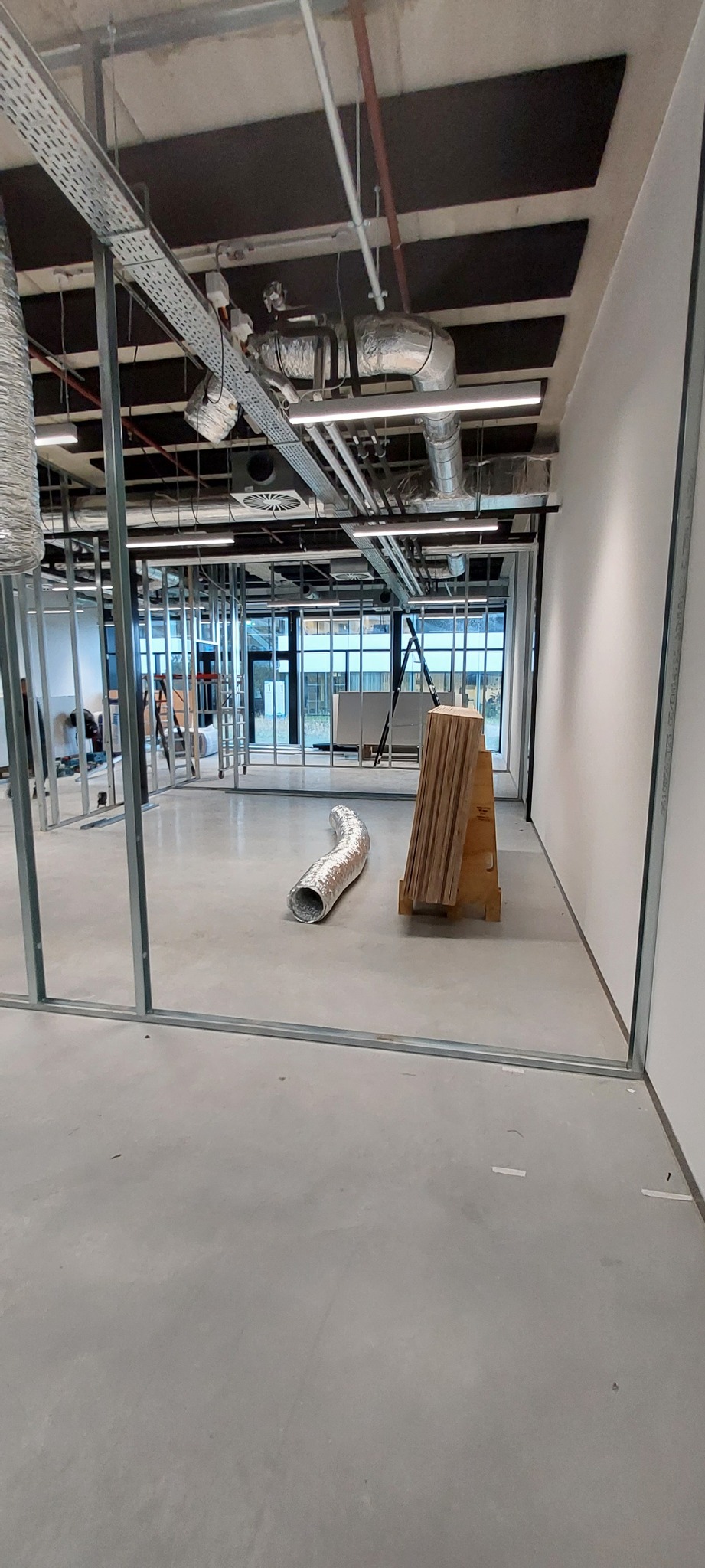 Stelaż metalowy do ścian działowych w surowym stanie, widoczny fragment wentylacji i składowane płyty, wnętrze biurowe w trakcie prac.