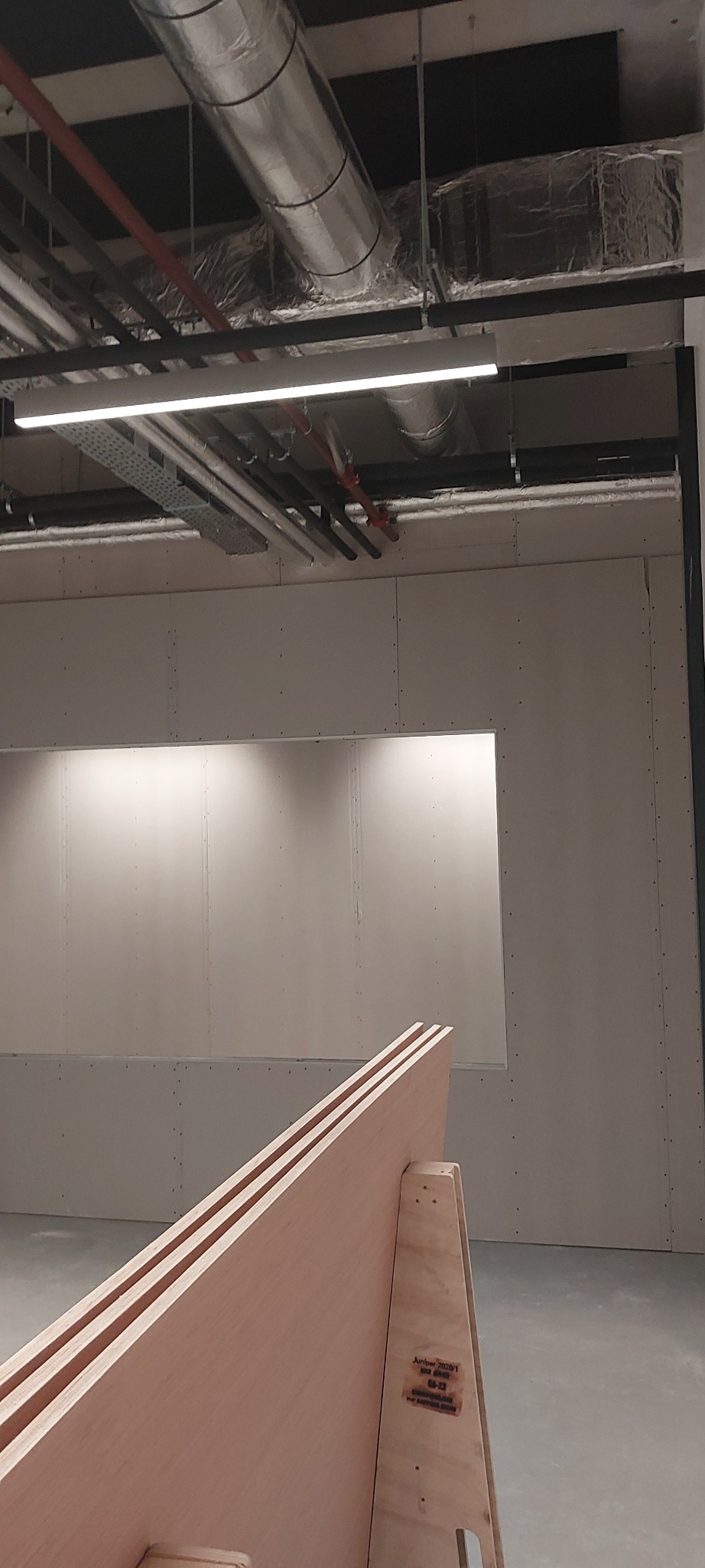 Wnętrze w trakcie prac wykończeniowych: ściany pokryte płytami gipsowo-kartonowymi z wyciętym otworem, widoczne instalacje pod sufitem i drewniany element konstrukcyjny na pierwszym planie.