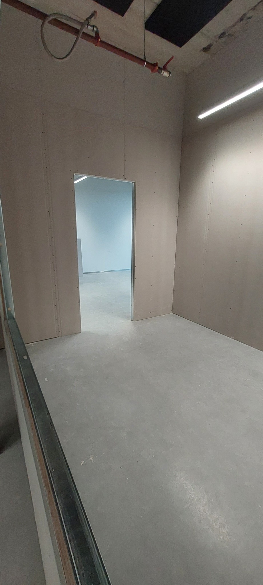 Surowe wnętrze: ściany wyłożone płytami gipsowo-kartonowymi, widoczna instalacja przeciwpożarowa pod sufitem, surowa podłoga i otwór drzwiowy do kolejnego pomieszczenia.