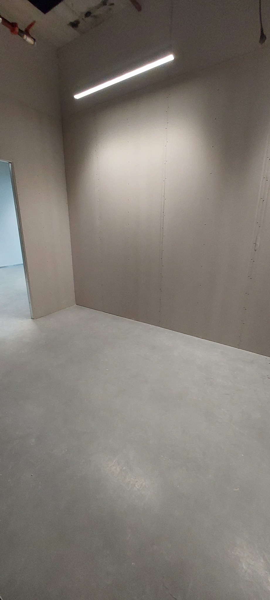 Surowe ściany z płyt, widoczne łączenia, szpachlowane, sufit z oświetleniem liniowym, podłoga betonowa, widok przez drzwi do kolejnego pomieszczenia.