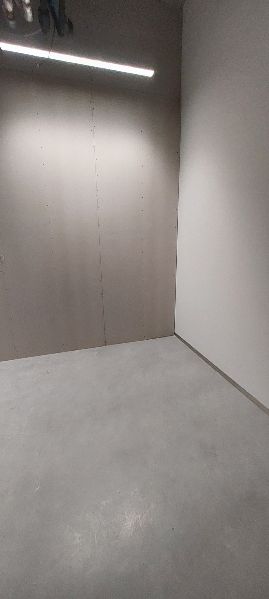 Wnętrze w trakcie prac wykończeniowych: ściana z zamontowaną płytą gipsowo-kartonową, widoczne łączenia płyt i wkręty, surowy betonowy sufit z lampą liniową, szara betonowa wylewka podłogowa.