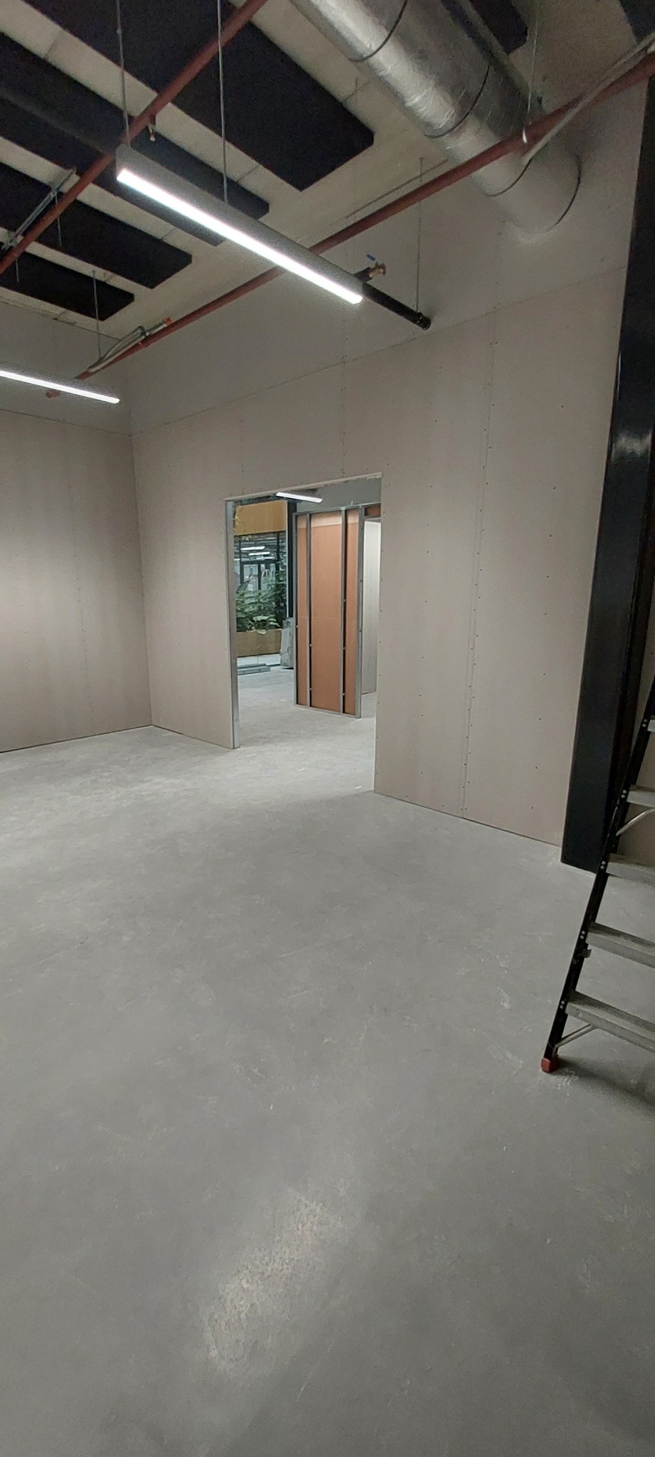 Wnętrze biurowe w trakcie remontu: ściany z płyt kartonowo-gipsowych, widoczne łączenia, surowy beton na podłodze, metalowa drabina w rogu, biuro w tle przez otwarte drzwi.