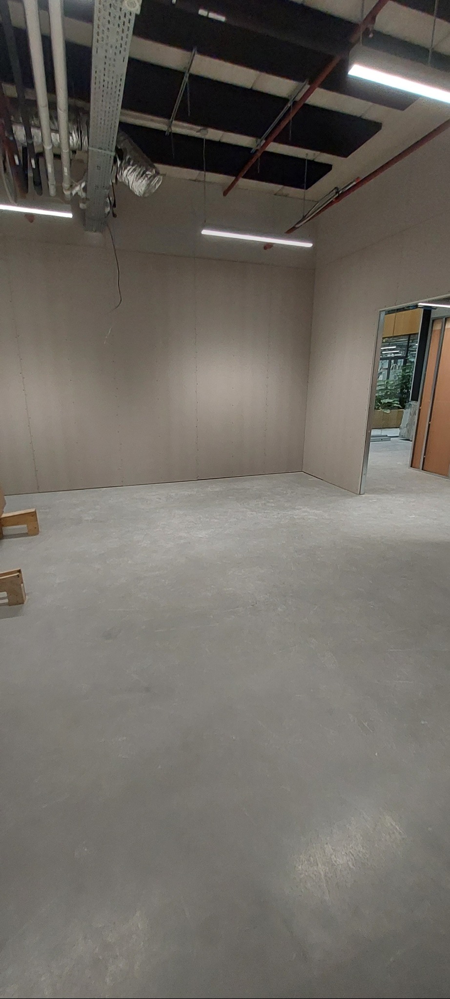 Surowe wnętrze biurowe z płytami na ścianach, widoczne rury wentylacyjne pod sufitem i otwarte przejście do kolejnego pomieszczenia.