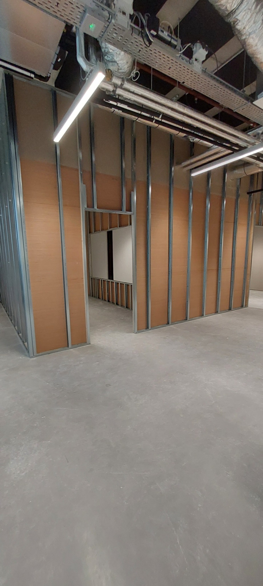 Stelaż z metalowych profili do montażu ścian działowych, częściowo obłożony płytami w kolorze jasnego drewna, widoczne instalacje pod sufitem.