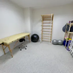 Jasne wnętrze gabinetu w Dąbrowie G&oacute;rniczej z beżowym stołem do masażu, drabinką gimnastyczną, piłkami do ćwiczeń i regałem z akcesoriami.