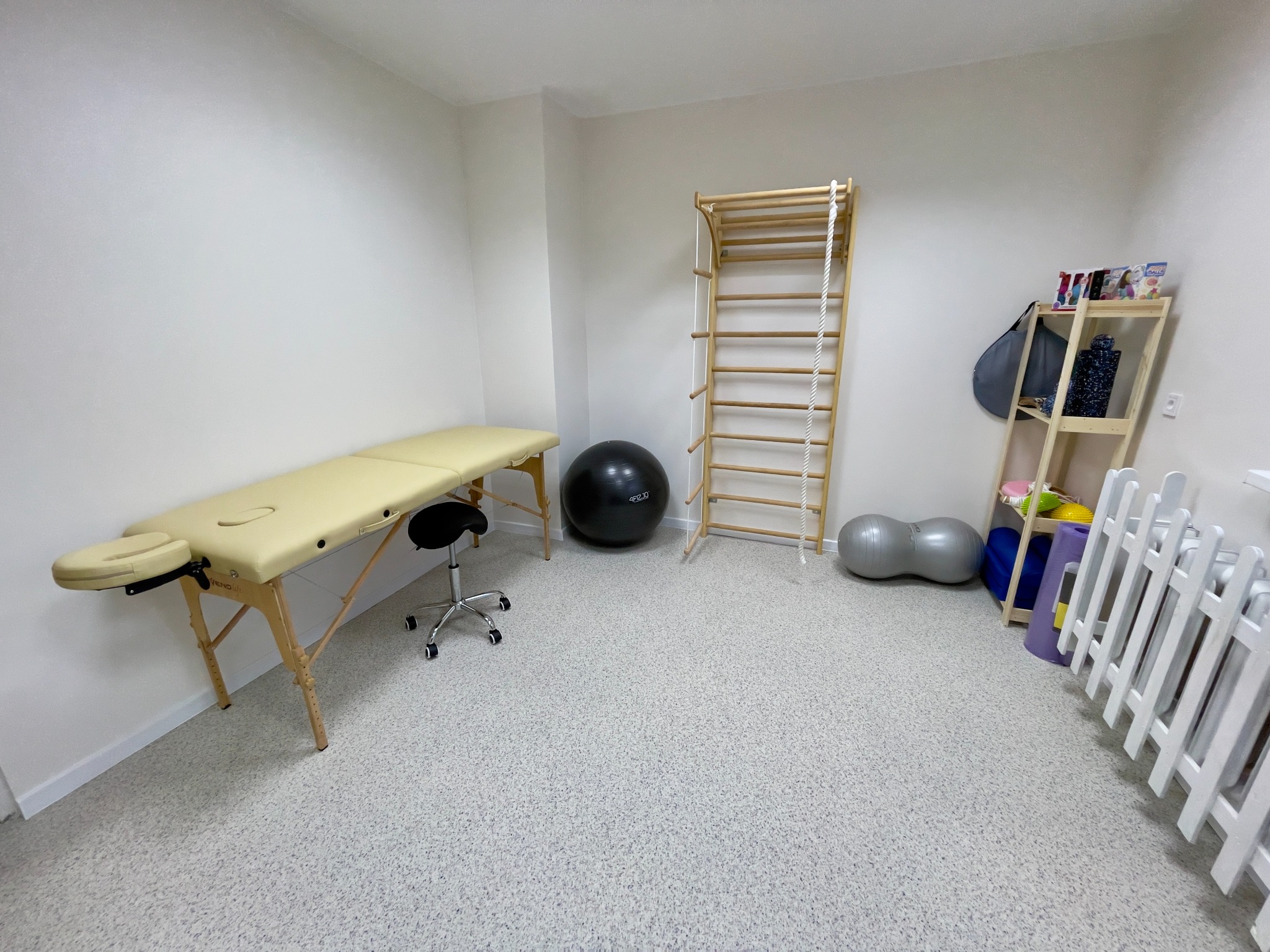 Jasne wnętrze gabinetu w Dąbrowie Górniczej z beżowym stołem do masażu, drabinką gimnastyczną, piłkami do ćwiczeń i regałem z akcesoriami.