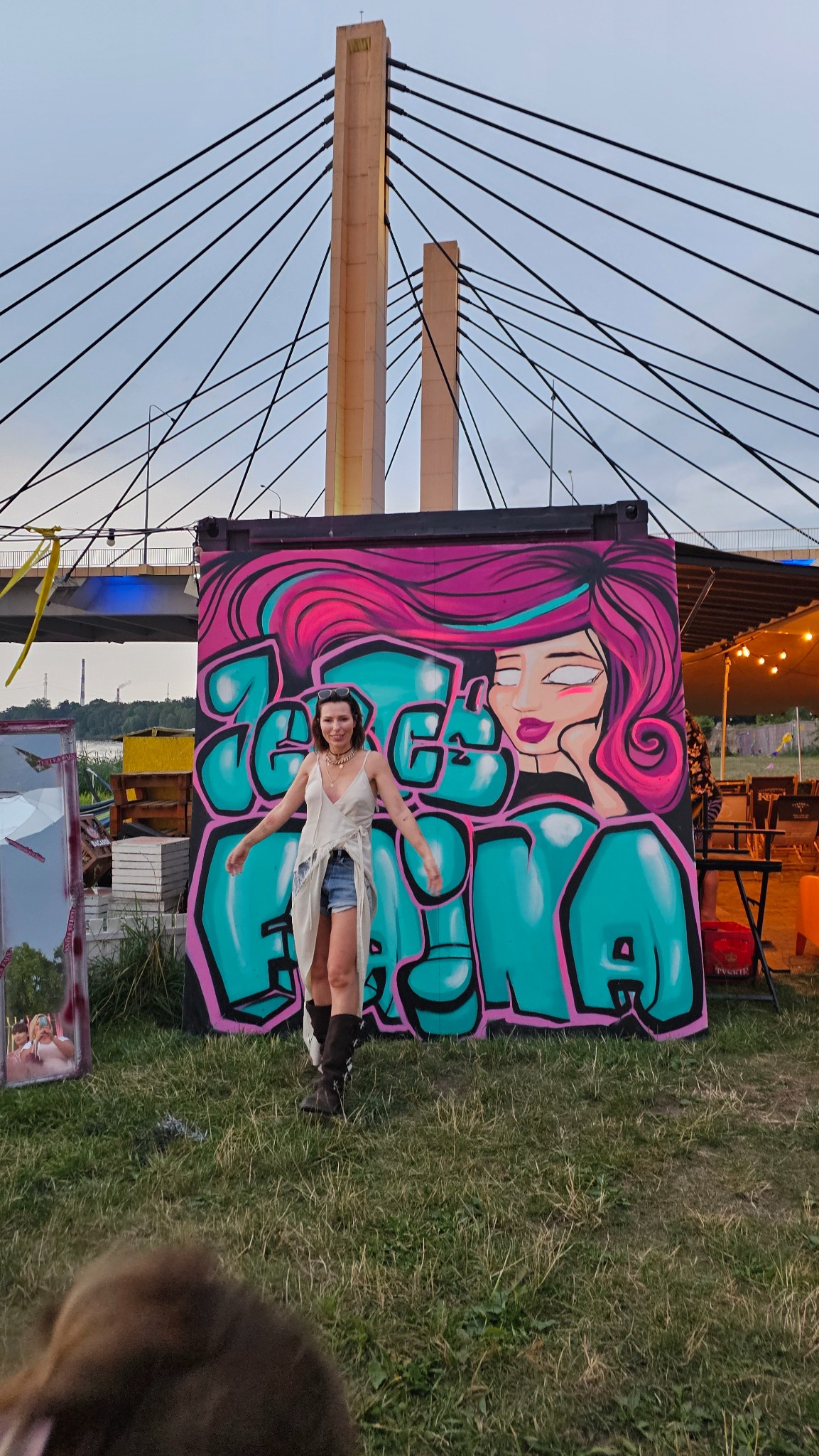 Kobieta pozuje przed pomalowanym kontenerem z graffiti przedstawiającym portret kobiety w stylu pop-art z różowymi włosami i turkusowymi napisami, w tle widoczny most z pylonami.
