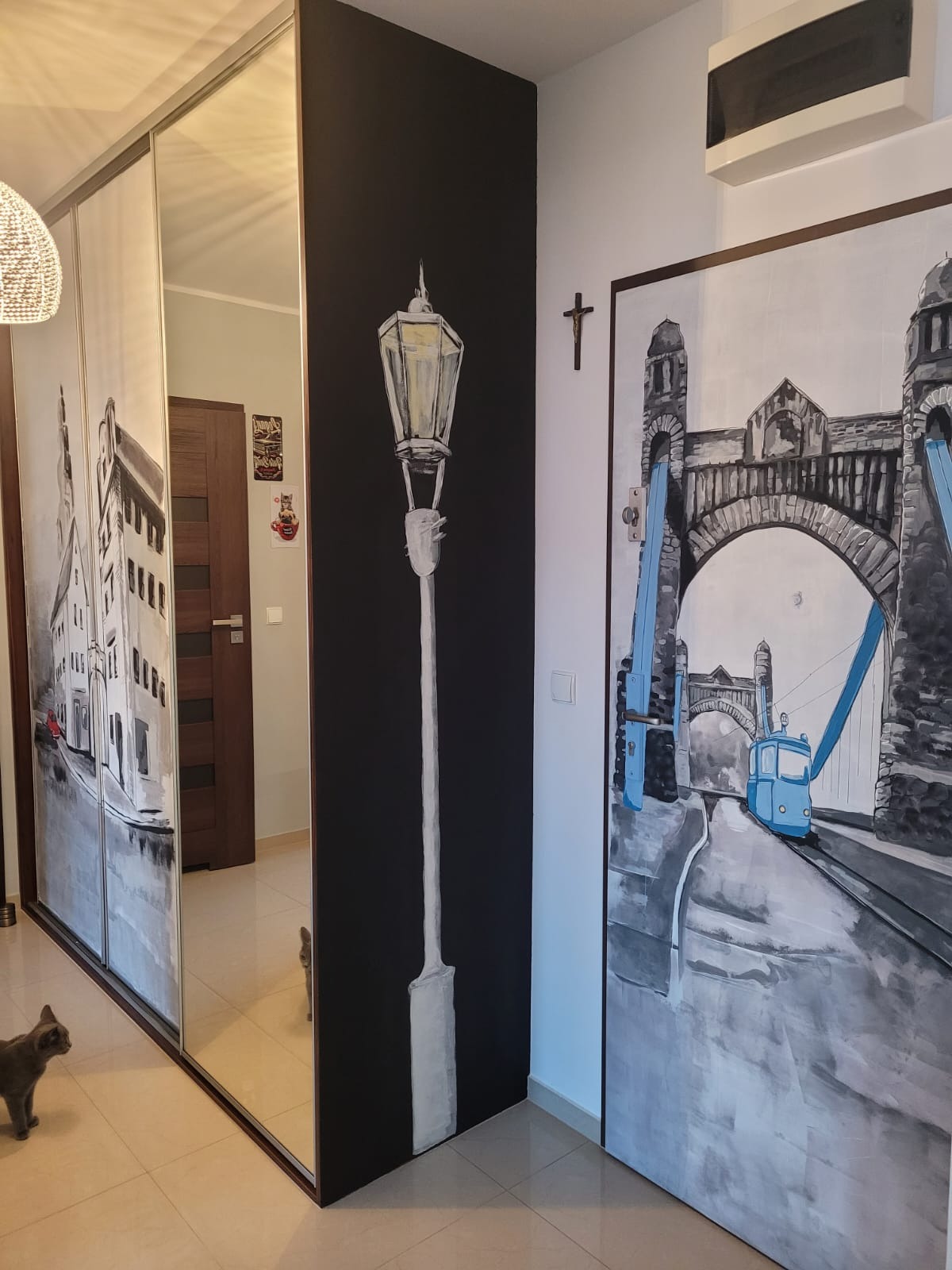 Wnętrze korytarza z ręcznie malowanymi obrazami na ścianach i szafie wnękowej: wizerunek wrocławskiej latarni, Mostu Grunwaldzkiego z tramwajem oraz kamienic. Szary kot w rogu kadru.