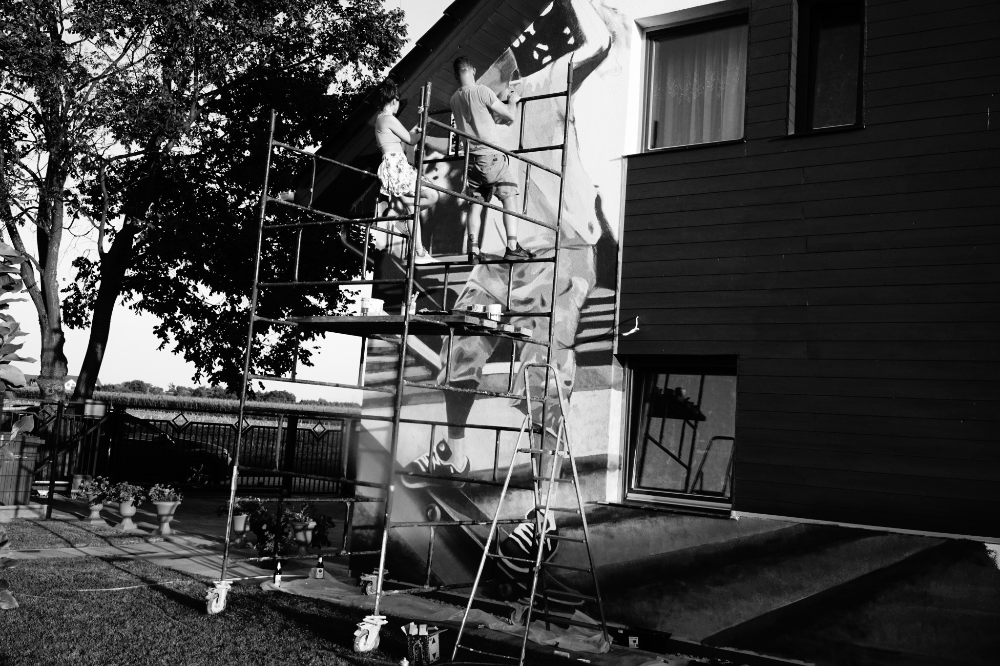Dwóch artystów na rusztowaniu maluje mural na ścianie domu; widoczne pędzle, drabina i donice z kwiatami.