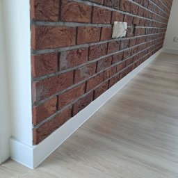 F.U.H GREGOR Grzegorz Mrozek - Ceglana ściana z fugą i białą listwą przypodłogową, oraz jasna podłoga imitująca drewno. Widoczny biały narożnik i gniazdka elektryczne.