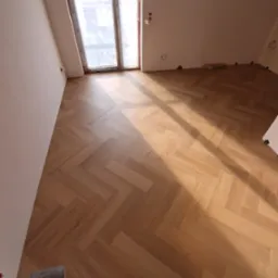 Ukończona podłoga z parkietu ułożonego w jodełkę w jasnym pokoju z oknem. Widoczne promienie słoneczne. Fragment narzędzia w lewym dolnym rogu.