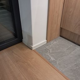 F.U.H GREGOR Grzegorz Mrozek - Połączenie paneli podłogowych z szarą, kamienną posadzką przy białej ścianie i czarnej ramie okiennej. Widoczne listwy przypodłogowe i wykończenie drewniane.