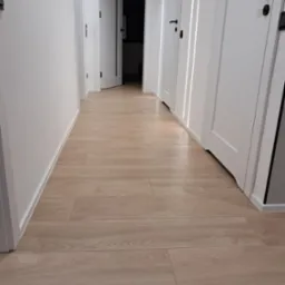 Jasne panele podłogowe w korytarzu z białymi ścianami i drzwiami z czarnymi klamkami. Widok z dołu podkreśla długość i perspektywę korytarza.