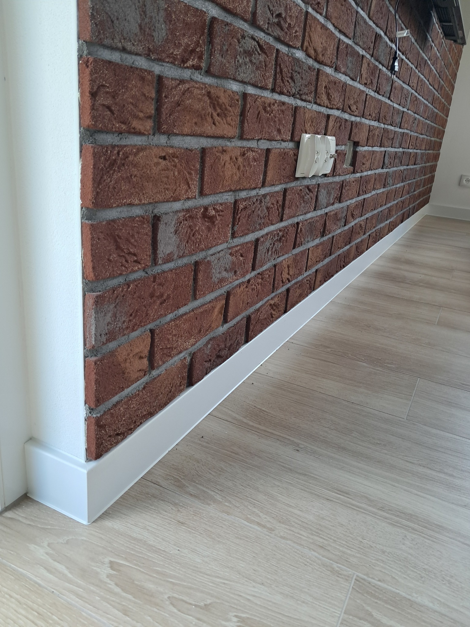 Ceglana ściana z fugą i białą listwą przypodłogową, oraz jasna podłoga imitująca drewno. Widoczny biały narożnik i gniazdka elektryczne.