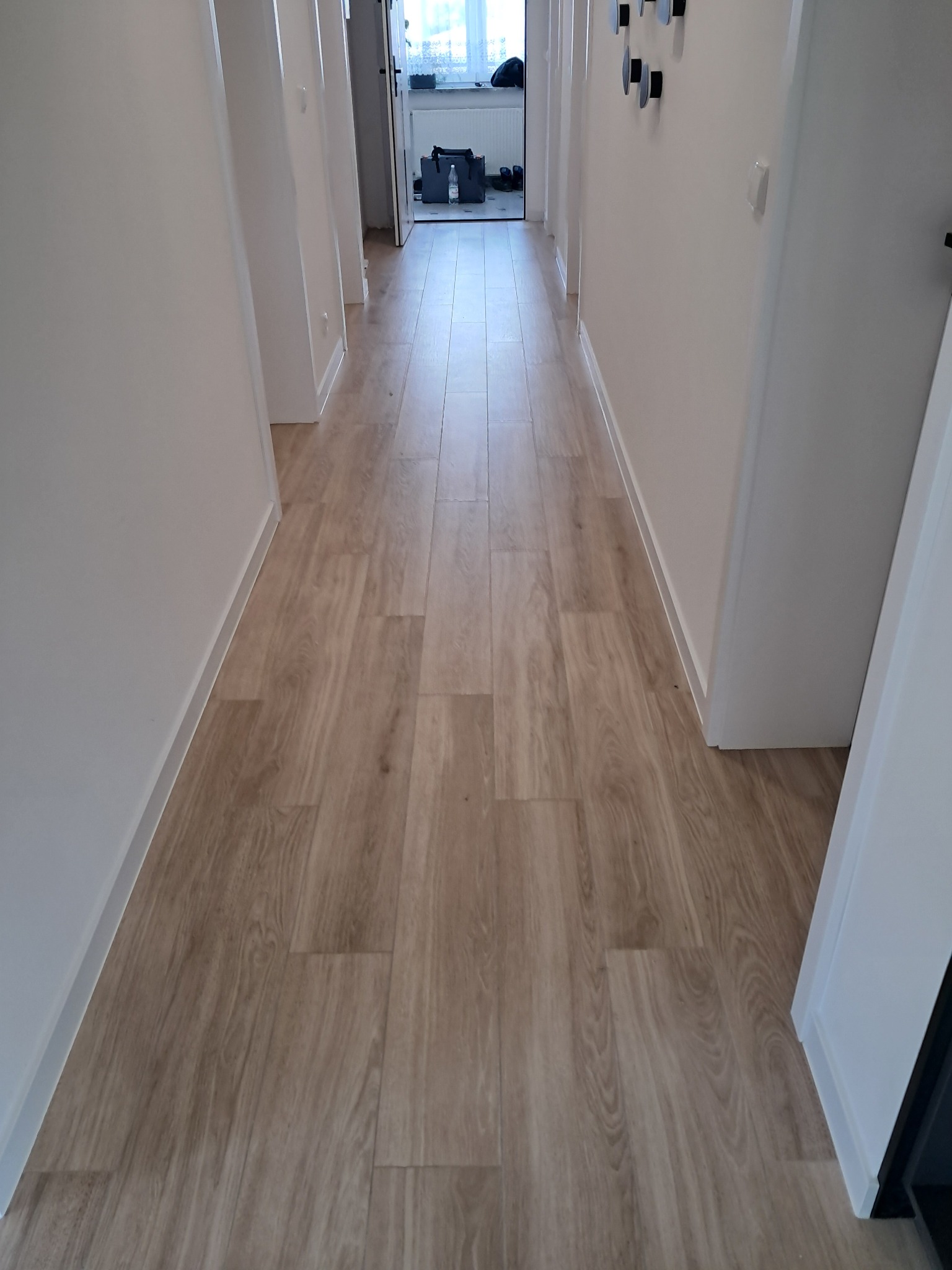 Długi korytarz z nową, jasną podłogą z paneli imitujących drewno. Białe ściany i drzwi. W tle widoczne okno i przedmioty.